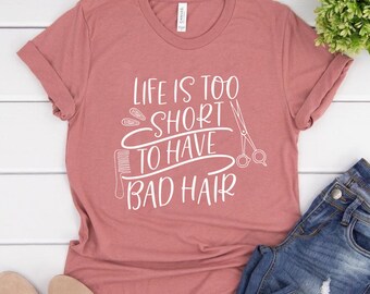 Puede incluir: Una camiseta de color rosa polvoriento con el texto "Life is too short to have bad hair" en blanco. El texto está rodeado de imágenes de herramientas para peinar el cabello, incluyendo tijeras, un peine y pinzas para el cabello.