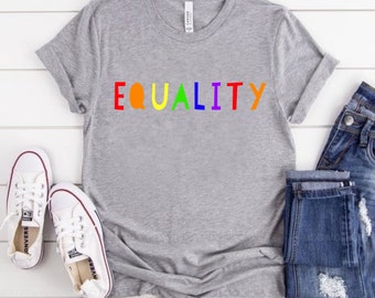 May include: A gray t-shirt with the word "EQUALITY" printed in rainbow colors. The shirt is folded and laid on a white wooden surface. A pair of white sneakers and a pair of blue jeans are also in the image.