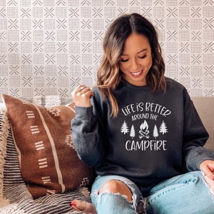 Puede incluir: Una sudadera gris oscura con el texto "Life is better around the campfire" en blanco. El texto est&aacute; rodeado por un gr&aacute;fico de una fogata con &aacute;rboles a cada lado.