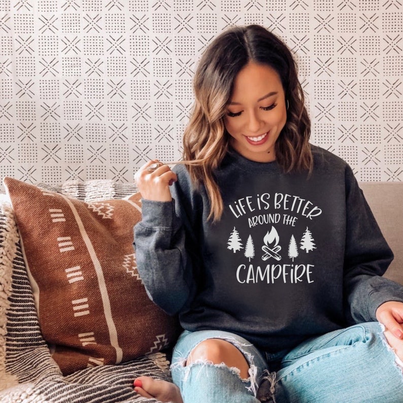 Puede incluir: Una sudadera gris oscura con el texto "Life is better around the campfire" en blanco. El texto est&aacute; rodeado por un gr&aacute;fico de una fogata con &aacute;rboles a cada lado.