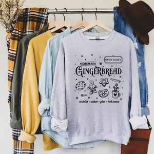 Może przedstawiać: Szara bluza z czarnym nadrukiem graficznym, na kt&oacute;rym widnieje napis "Homemade Gingerbread" wraz z piernikowym ludzikiem, ciasteczkami, preclami i kubkiem gorącej czekolady. Tekst pod grafiką brzmi "cookies - cakes - pies - hot cocoa" i "Open Daily".