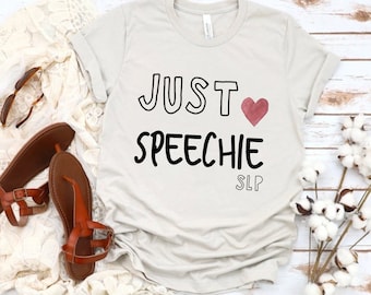 Puede incluir: Una camiseta gris claro con el texto "JUST ♥ SPEECHIE SLP" impreso en negro. El corazón es rosa.