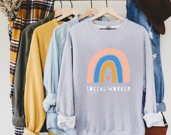 May include: A gray sweatshirt with a rainbow design and the text "SOCIAL WORKER" in white. The rainbow is made up of pink, blue, and yellow stripes with a small red heart in the center.
