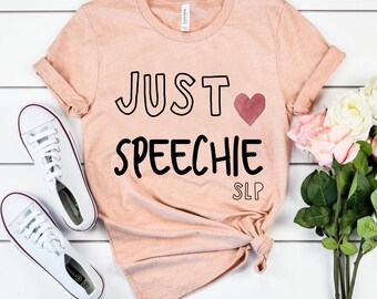 Puede incluir: Una camiseta de color melocotón con el texto "JUST ♥ SPEECHIE SLP" impreso en negro.