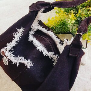May include: A black denim shoulder bag with white fringe trim and a zipper closure. The bag has a long strap and a metal buckle.