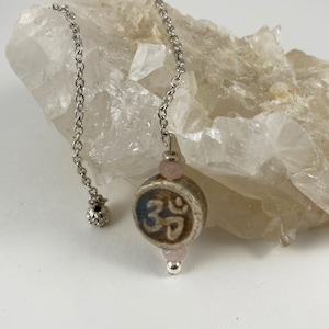 May include: A silver chain with a brown and blue ceramic pendant featuring an Om symbol. The pendant is accented with two small pink beads.