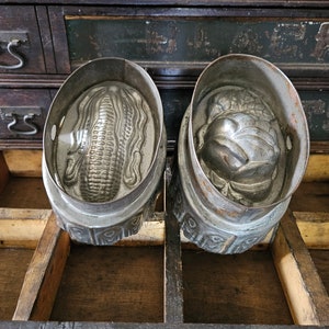 May include: Two vintage metal cake molds, one shaped like an ear of corn and the other shaped like a bird, sitting in a wooden drawer.