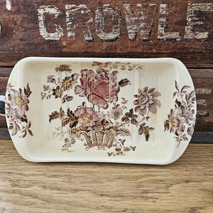 May include: Cream-colored rectangular soap dish with a floral design in shades of brown and pink. The soap dish features a central floral arrangement in a basket, with additional floral patterns on the sides. The dish has rounded edges and a slightly raised rim.