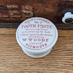 May include: A white round tin with red lettering that reads "WOODS CHERRY 6d TOOTH PASTE Forremoving Tartar and whitening the Teeth without injuring the Enamel PROPRIETOR W. WOODS CHEMIST PLYMOUTH".