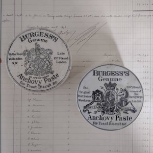 May include: Two vintage, black and white, round, tin containers for anchovy paste. The containers have a royal crest design and the text "Burgess's Genuine Anchovy Paste for Toast Biscuit &c."  The text on the left container reads "Hythe Road Willesden N.W. Late 107 Strand London." The text on the right container reads "The Original Fish Sauce Warehouse 107 Strand corner of the Savoy Steps London."