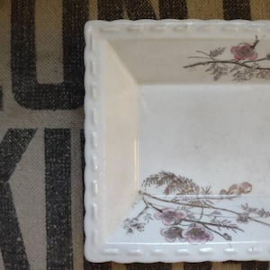 May include: A rectangular, white ceramic dish with a scalloped edge. The dish is decorated with a floral design featuring pink blossoms and brown branches. The dish is likely used for serving or display.