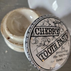 May include: An antique ceramic jar with a lid featuring the words "CHERRY TOOTH PASTE" in a vintage font. The lid has a black and white cherry illustration and text that reads "for preserving the teeth and gums."