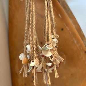 May include: A macrame seashell hanging decoration. The natural fiber rope is knotted to hold various seashells. The shells are white, tan, and cream-colored. The hanging decoration is held up by a hand.