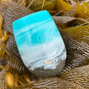 May include: A stemless tumbler with a coastal design. The top is a bright turquoise, transitioning to white and gray, resembling ocean waves. The base is a sandy color. The tumbler rests on brown seaweed.