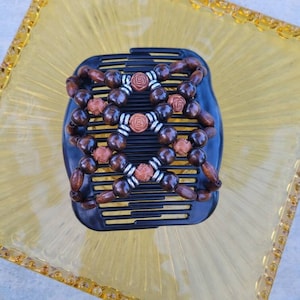 May include: A black hair comb with wooden beads and copper-coloured accents. The dark brown beads are arranged in a symmetrical pattern. The comb is set on a yellow, textured surface.