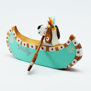May include: A wooden toy canoe with a dog wearing a headdress paddling. The canoe is painted teal with red and white accents. The dog is white with brown spots and the name "Willy" is written on the side of the canoe.