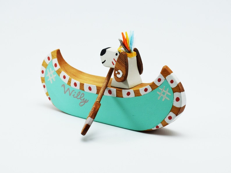 May include: A wooden toy canoe with a dog wearing a headdress paddling. The canoe is painted teal with red and white accents. The dog is white with brown spots and the name "Willy" is written on the side of the canoe.