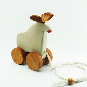 May include: A white wooden pull toy shaped like a moose with brown antlers and a red nose. The toy has four wooden wheels and a white rope attached to the back.