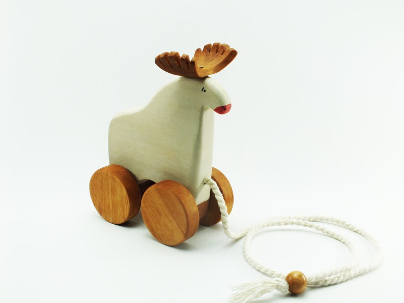 May include: A white wooden pull toy shaped like a moose with brown antlers and a red nose. The toy has four wooden wheels and a white rope attached to the back.