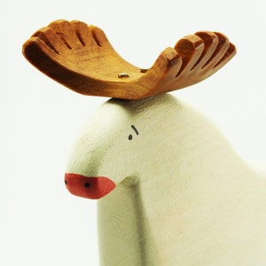 May include: A white wooden moose figurine with a brown wooden antler. The moose has a red painted nose and black painted eyes.