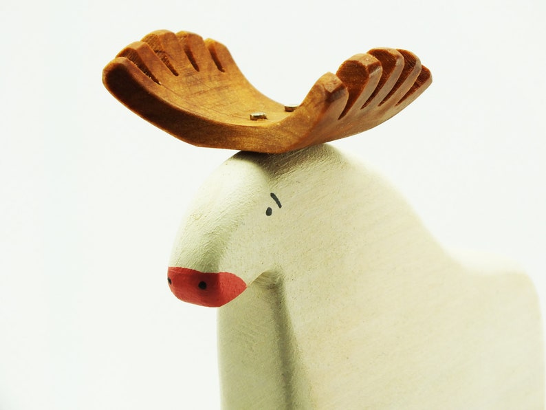 May include: A white wooden moose figurine with a brown wooden antler. The moose has a red painted nose and black painted eyes.
