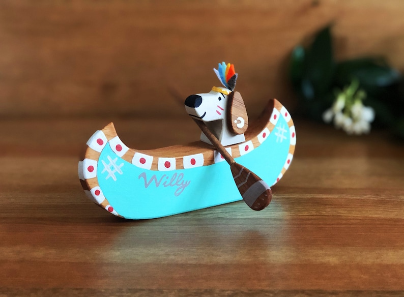 May include: A wooden toy canoe painted turquoise with red and white accents. A small wooden dog figure with a feathered headdress is paddling the canoe. The canoe is labeled "Willy".