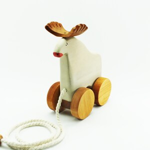 May include: A white wooden pull toy shaped like a moose with brown antlers and wheels. The toy has a red painted nose and a white rope attached to it.