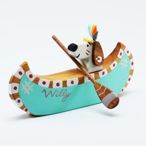 May include: A wooden toy canoe painted turquoise with red and white accents. A white dog with a brown head and a colorful feather headdress is paddling the canoe. The canoe is labeled "Willy".