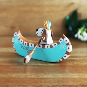 May include: A wooden canoe with a teal blue hull and red and white accents. The canoe is decorated with a white snowflake and the name "Willy". A small wooden dog figure with a colorful feather headdress is sitting in the canoe, holding a wooden paddle.