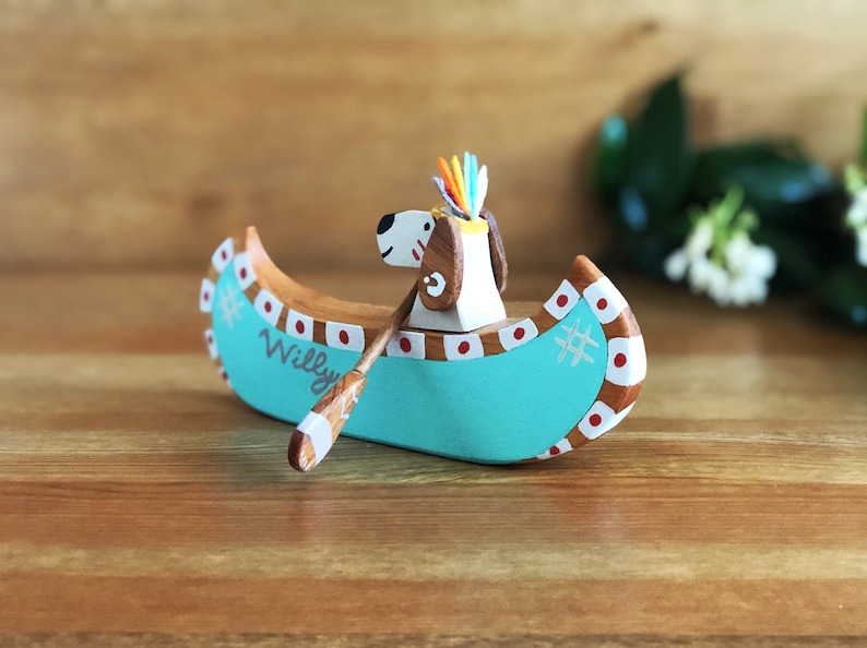 May include: A wooden canoe with a teal blue hull and red and white accents. The canoe is decorated with a white snowflake and the name "Willy". A small wooden dog figure with a colorful feather headdress is sitting in the canoe, holding a wooden paddle.