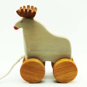May include: A white wooden pull toy shaped like a reindeer with brown antlers and red painted nose. The toy has four wooden wheels and a white string attached to the back.
