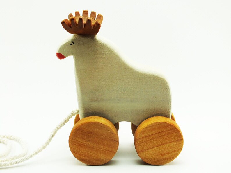 May include: A white wooden pull toy shaped like a reindeer with brown antlers and red painted nose. The toy has four wooden wheels and a white string attached to the back.