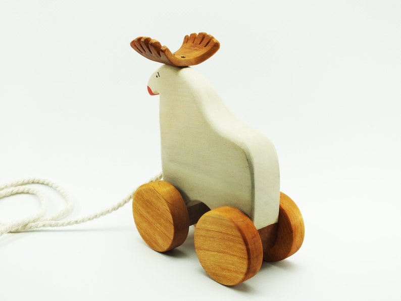 May include: A white wooden pull toy shaped like a moose with brown wooden antlers and wheels. The toy has a string attached to it.
