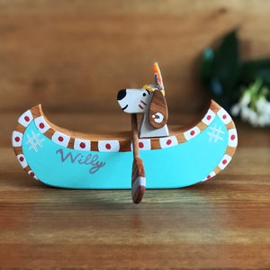 May include: A wooden toy canoe with a dog figure paddling. The canoe is painted turquoise with red and white accents. The dog is white with brown spots and has a red and yellow headdress. The canoe is personalized with the name "Willy".
