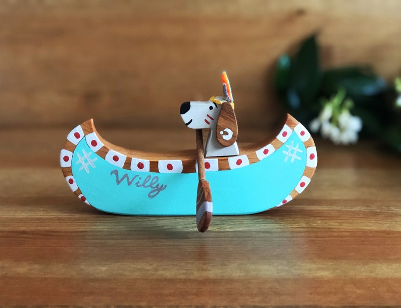 May include: A wooden toy canoe with a dog figure paddling. The canoe is painted turquoise with red and white accents. The dog is white with brown spots and has a red and yellow headdress. The canoe is personalized with the name "Willy".