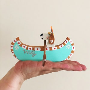 May include: A wooden toy canoe with a dog's head at the bow. The canoe is painted turquoise with red and white accents. The canoe is labeled "Willy".