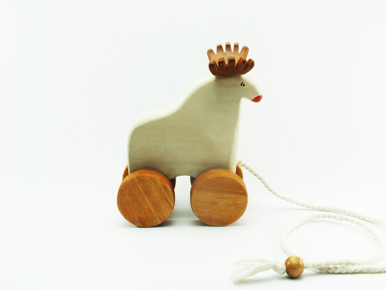 May include: A white wooden pull toy shaped like a reindeer with brown antlers and red painted lips. The toy has two wooden wheels and a white string attached to the back.