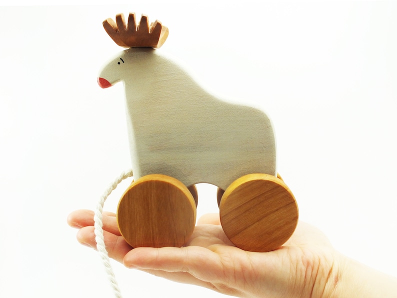 May include: A white wooden toy reindeer with brown antlers and red nose on wheels. The toy is being held in a hand.