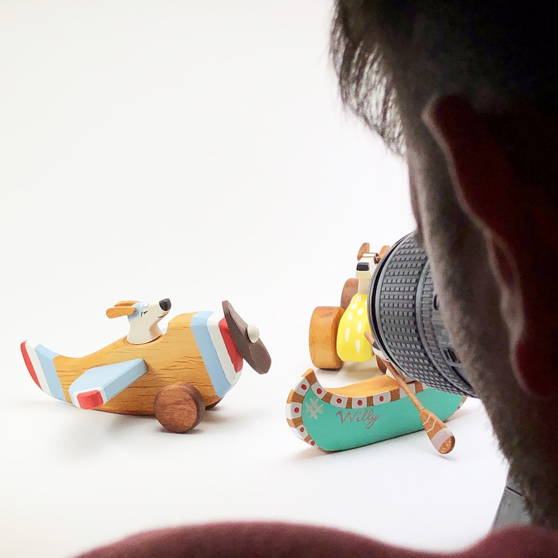 May include: A set of three wooden toys, including a blue and white airplane with a dog pilot, a yellow and red car with a driver, and a teal canoe with the word "Willy" on it.