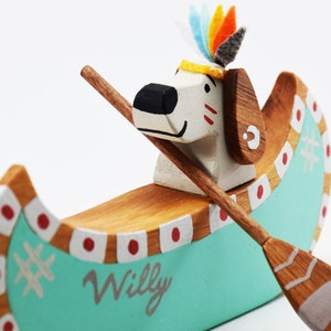 May include: A wooden toy canoe with a dog figure paddling. The canoe is painted teal with white and red accents. The dog is wearing a colorful headdress and has a painted face. The canoe is labeled "Willy".