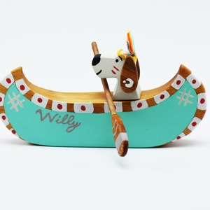 May include: A wooden toy canoe with a dog paddling. The canoe is painted teal blue with red and white accents. The dog is white with brown ears and a red and white headdress. The canoe is labeled "Willy".
