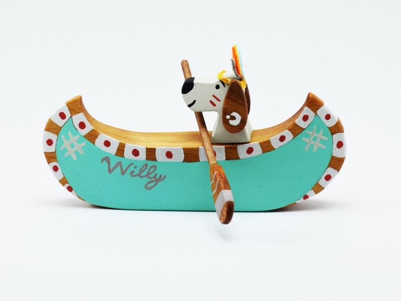 May include: A wooden toy canoe with a dog paddling. The canoe is painted teal blue with red and white accents. The dog is white with brown ears and a red and white headdress. The canoe is labeled "Willy".