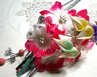 Puede incluir: Un adorno tradicional para el cabello japonés con un grupo de flores de seda de colores. Las flores están hechas de seda blanca, rosa, amarilla y verde y están decoradas con detalles dorados y plateados. El adorno está unido a un par de palillos de plata.