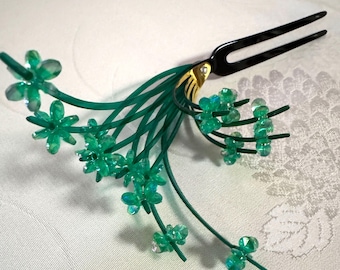 May include: A decorative hair accessory with a black two-pronged comb and gold detailing. Green stems with clear, faceted flower-shaped beads extend from the comb, forming a floral design. The accessory is set against a white background.