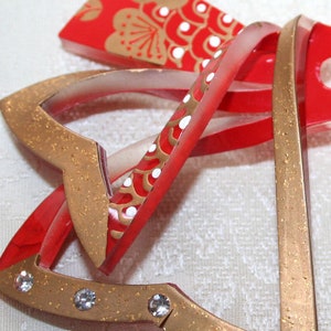 May include: A set of three gold and red Christmas bells with white accents and rhinestones. The bells are layered on top of each other and have a sparkly finish.