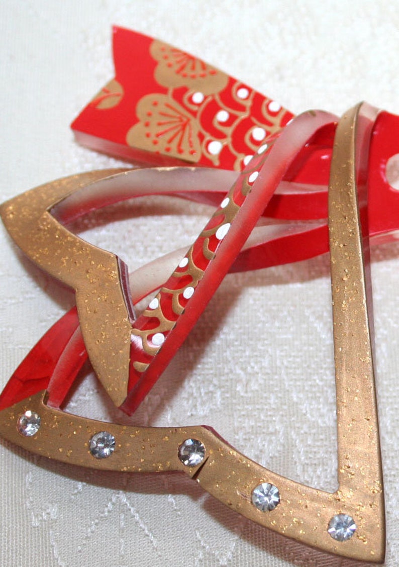 May include: A set of three gold and red Christmas bells with white accents and rhinestones. The bells are layered on top of each other and have a sparkly finish.