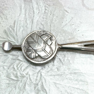 May include: A silver-toned metal hair accessory featuring a circular centerpiece with a textured background and a raised, polished cross-like design composed of four leaf-shaped segments. The piece includes a thin, straight stem with a rounded end.