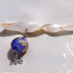 May include: A white, twisted, shell hair stick with a purple and gold fabric ball charm hanging from it. The charm has a silver chain and a silver bead tassel.