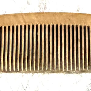 May include: A light brown, rectangular hair comb featuring a series of thin, evenly spaced vertical teeth. The top edge of the comb is slightly arched, and the material displays a natural, textured appearance with subtle variations in color.