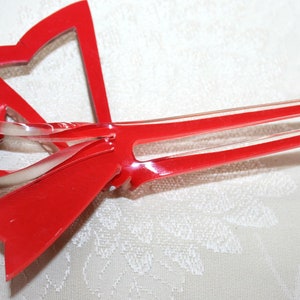 May include: A red plastic hair clip shaped like a bow with two prongs. The bow is translucent and has a glossy finish.